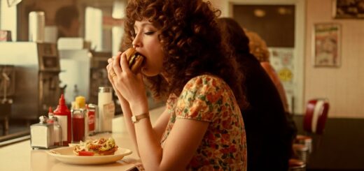 Eine Frau mit welligem Haar sitzt an einem Tresen in einem Diner der 70er Jahre und genießt einen Burger.