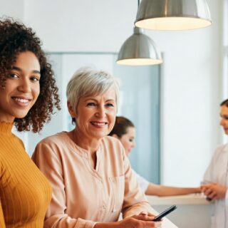 Zwei Frauen, eine jüngere und eine ältere, stehen mit Smartphones in der Hand an einem hellen Empfangstresen in einer Arztpraxis. Zwei Arzthelferinnen in weißen Kitteln stehen hinter dem Tresen.