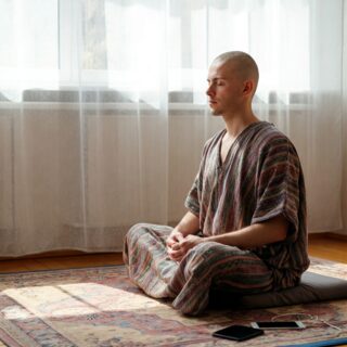 Das Bild zeigt eine entspannte Person, die in einem ruhigen, lichtdurchfluteten Raum sitzt und meditiert. Im Hintergrund sind nur leise andeutungsweise technische Geräte zu sehen, die ausgeschaltet sind, um den Fokus auf den Zustand der inneren Balance und Erholung zu legen. Das Bild vermittelt eine realistische, entspannte Atmosphäre, passend zum Thema Digital Detox.