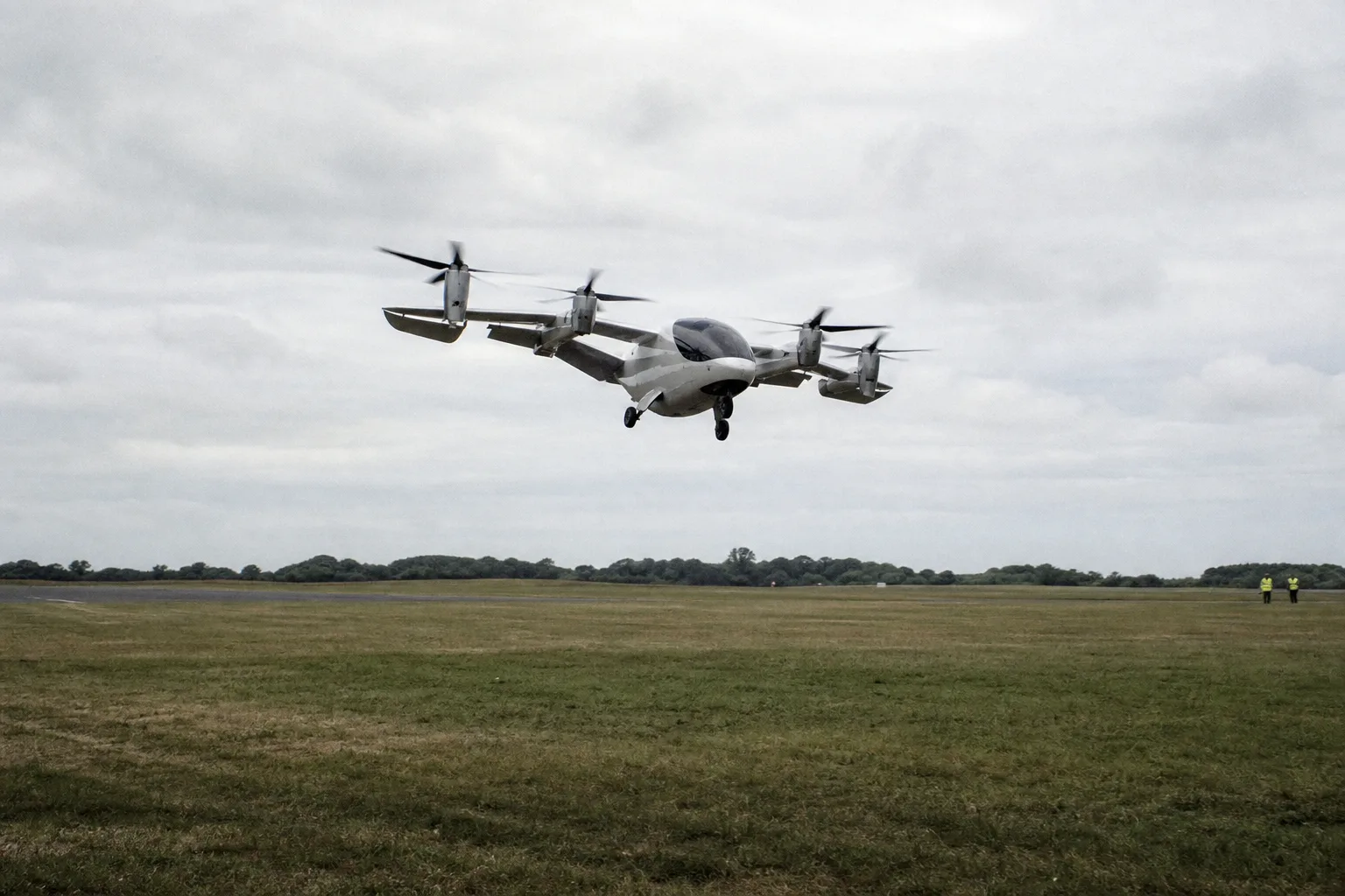 Ein unmarkiertes elektrisch angetriebenes Senkrechtstartflugzeug geht über einer breiten Grasbahn von einem steilen Steigflug in eine flachere Fluglage über; im Hintergrund ist ein Flugplatz zu sehen.