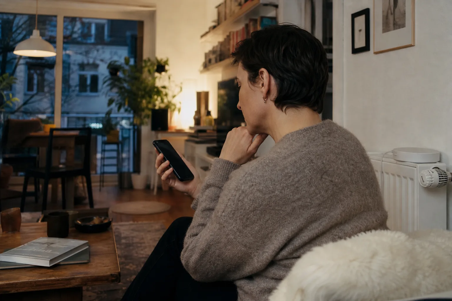 Hand hält ein Smartphone in einem Wohnzimmer neben einem neutralen Heizkörperthermostat; die Anzeige ist nicht lesbar.