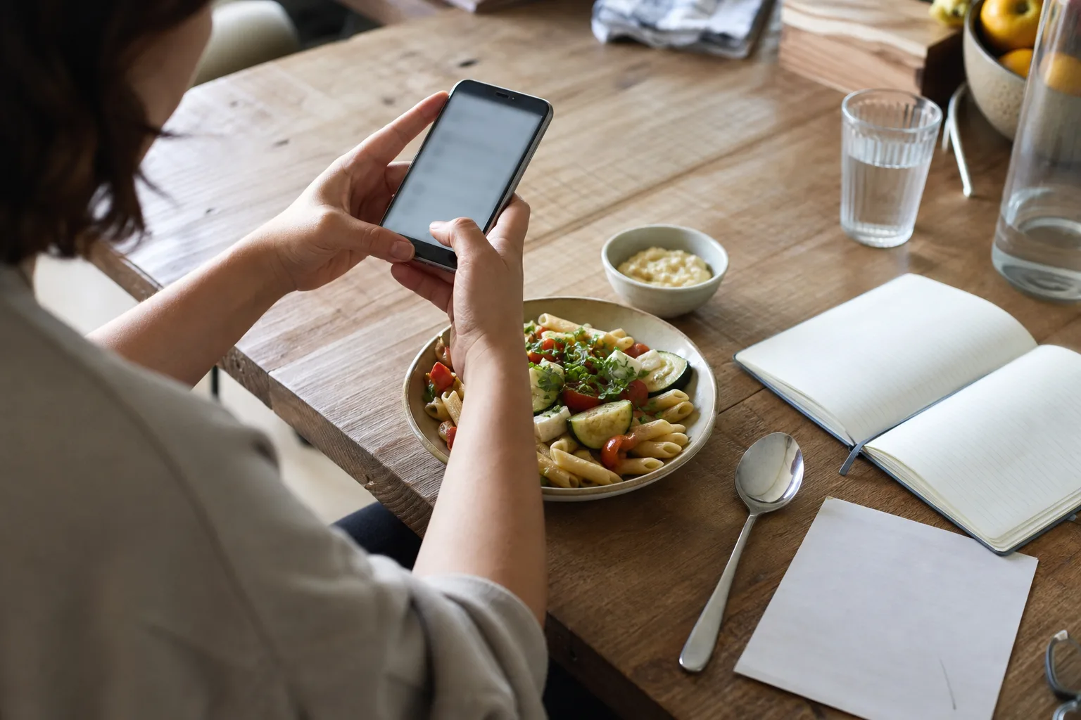 Eine Person fotografiert an einem Küchentisch eine Mahlzeit mit Pasta und Gemüse; das Smartphone-Display ist unscharf.