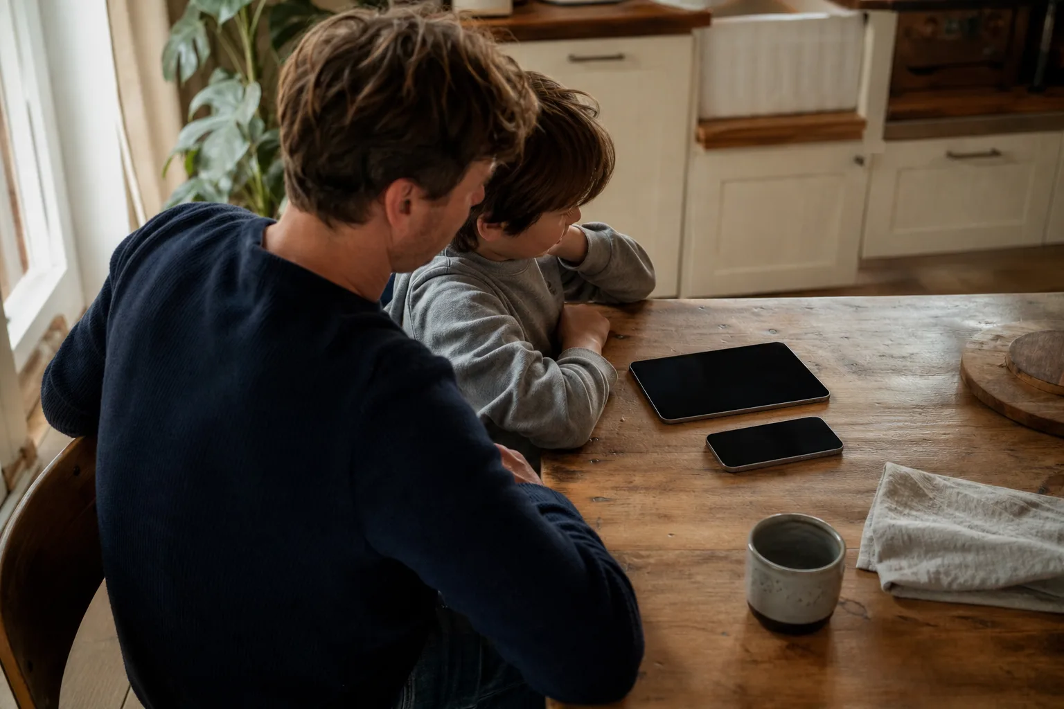 Ein Elternteil und ein Kind prüfen gemeinsam ein neutrales Smartphone und Tablet am Küchentisch.