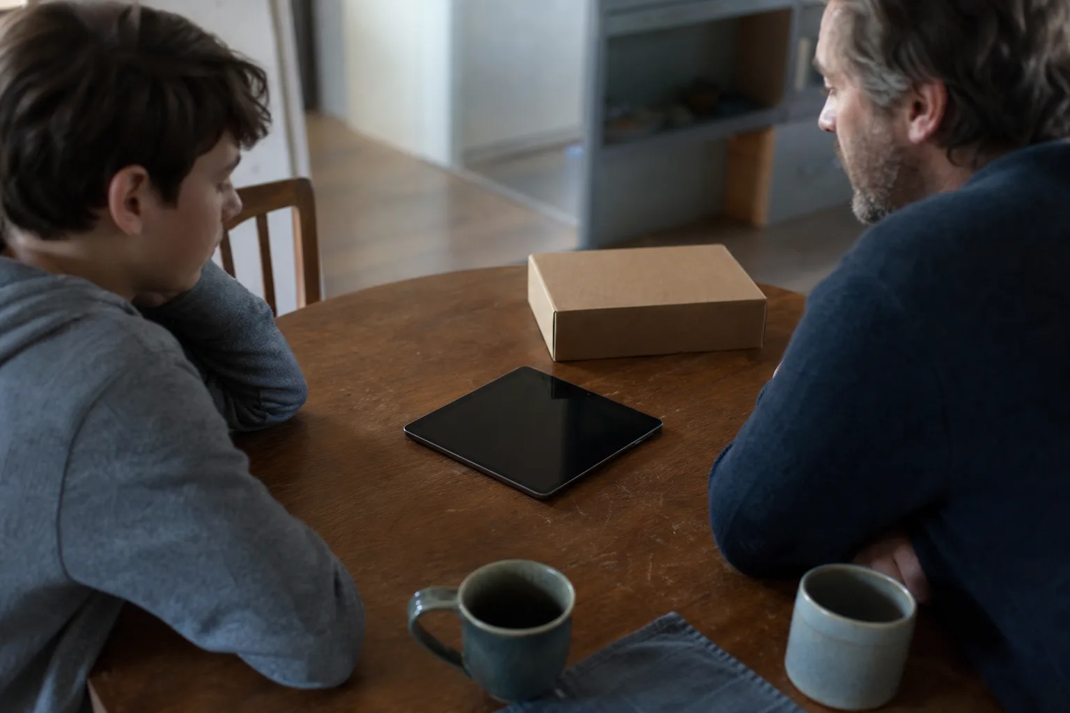 Zwei Familienmitglieder besprechen am Küchentisch Regeln neben einem neutralen Tablet mit schwarzem Display.