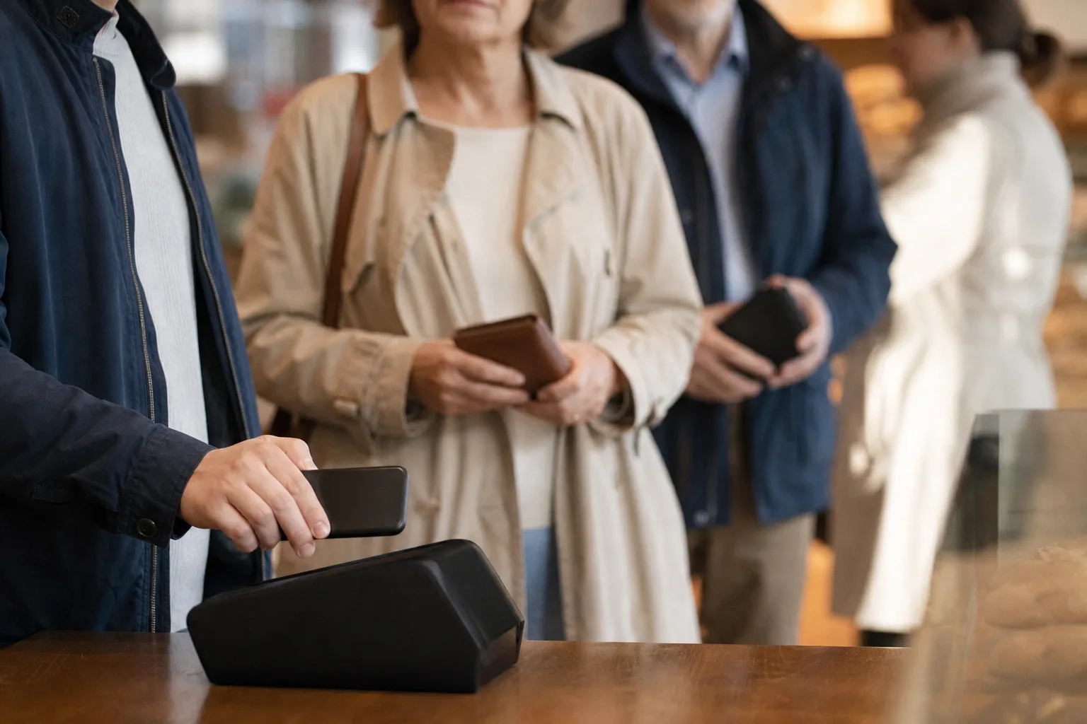 Eine Person bezahlt mit einem Smartphone, zwei weitere warten mit geschlossenen Portemonnaies an einer Ladenkasse.