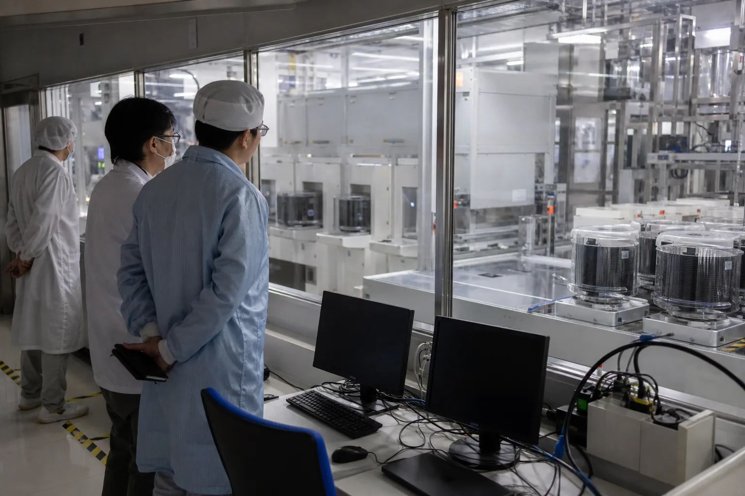 Ingenieure beobachten in einer modernen Halbleiterfertigung Anlagen für Speicherchips hinter einer Glasscheibe.