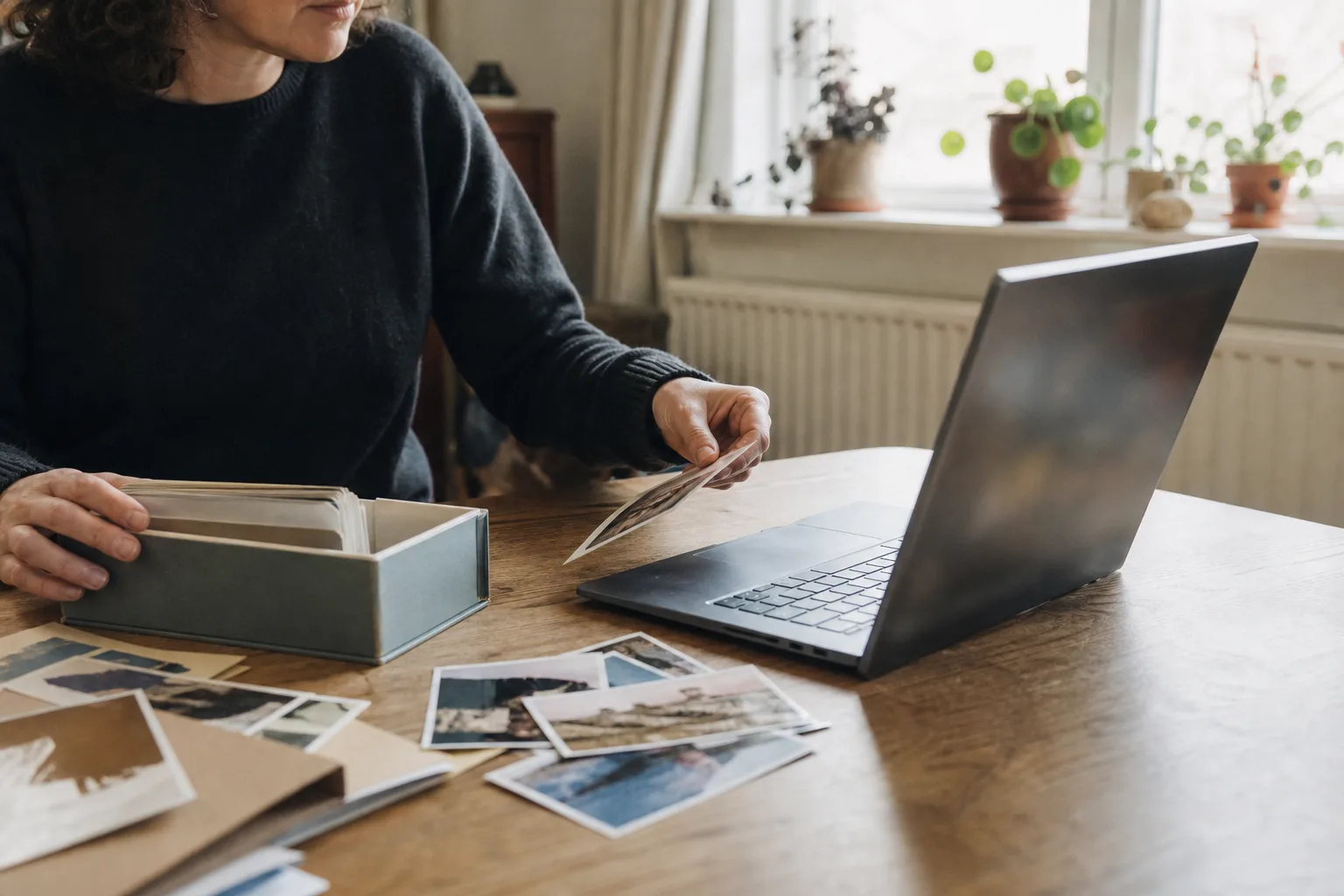 Person sortiert zuhause private Fotoabzüge neben einem neutralen Laptop und einem Ordner mit Familienbildern.