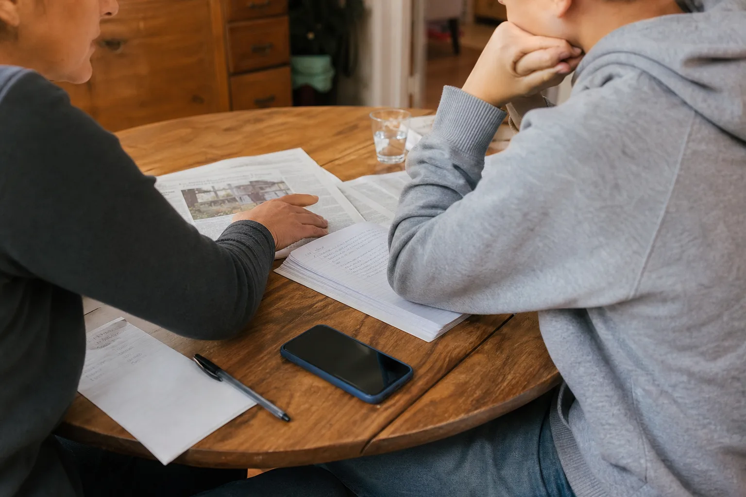 Elternteil und Jugendlicher besprechen an einem Küchentisch eine gedruckte Nachrichtenseite neben einem beiseitegelegten Smartphone