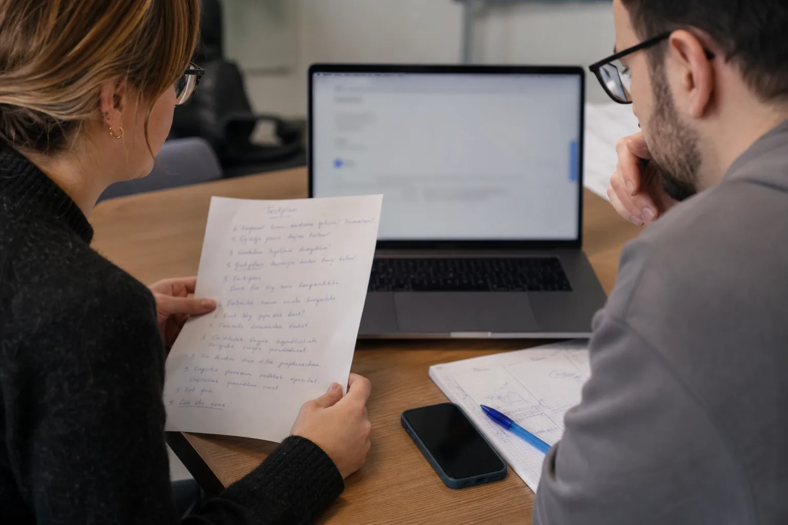 Zwei Personen prüfen gemeinsam an Laptop und Smartphone den Kernablauf einer Anwendung mit Testnotizen auf dem Tisch.