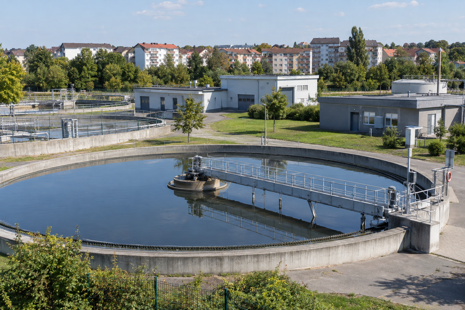 Nachklärbecken eines kommunalen Klärwerks mit Betriebsgebäuden und räumlich getrennter Wohnbebauung im Hintergrund.